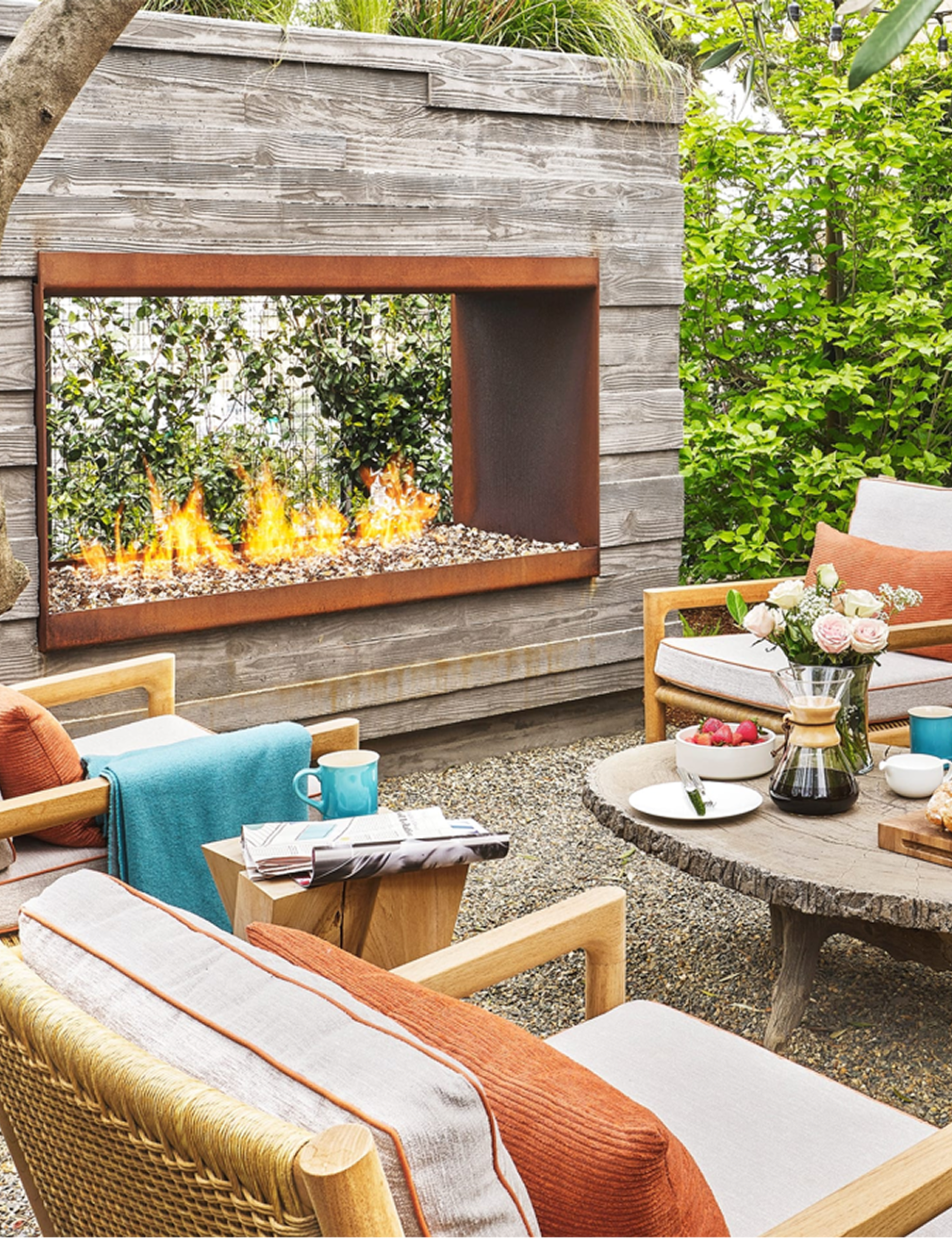 Warm, rustic outdoor patio area featuring a double-sided fire feature and a cushioned seating group punctuated by teal throw blankets and mugs.