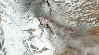 An image from space showing a gray plume of smoke drifting over brown and snowy mountains