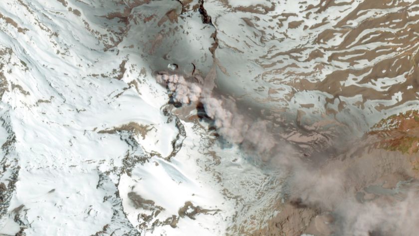 An image from space showing a gray plume of smoke drifting over brown and snowy mountains