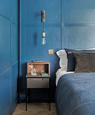 Bedroom with blue panelled walls, double bed with blue denim bedding and blue and black cushions, wall lamp with exposed hanging light bulb, wooden bedside table and wooden flooring