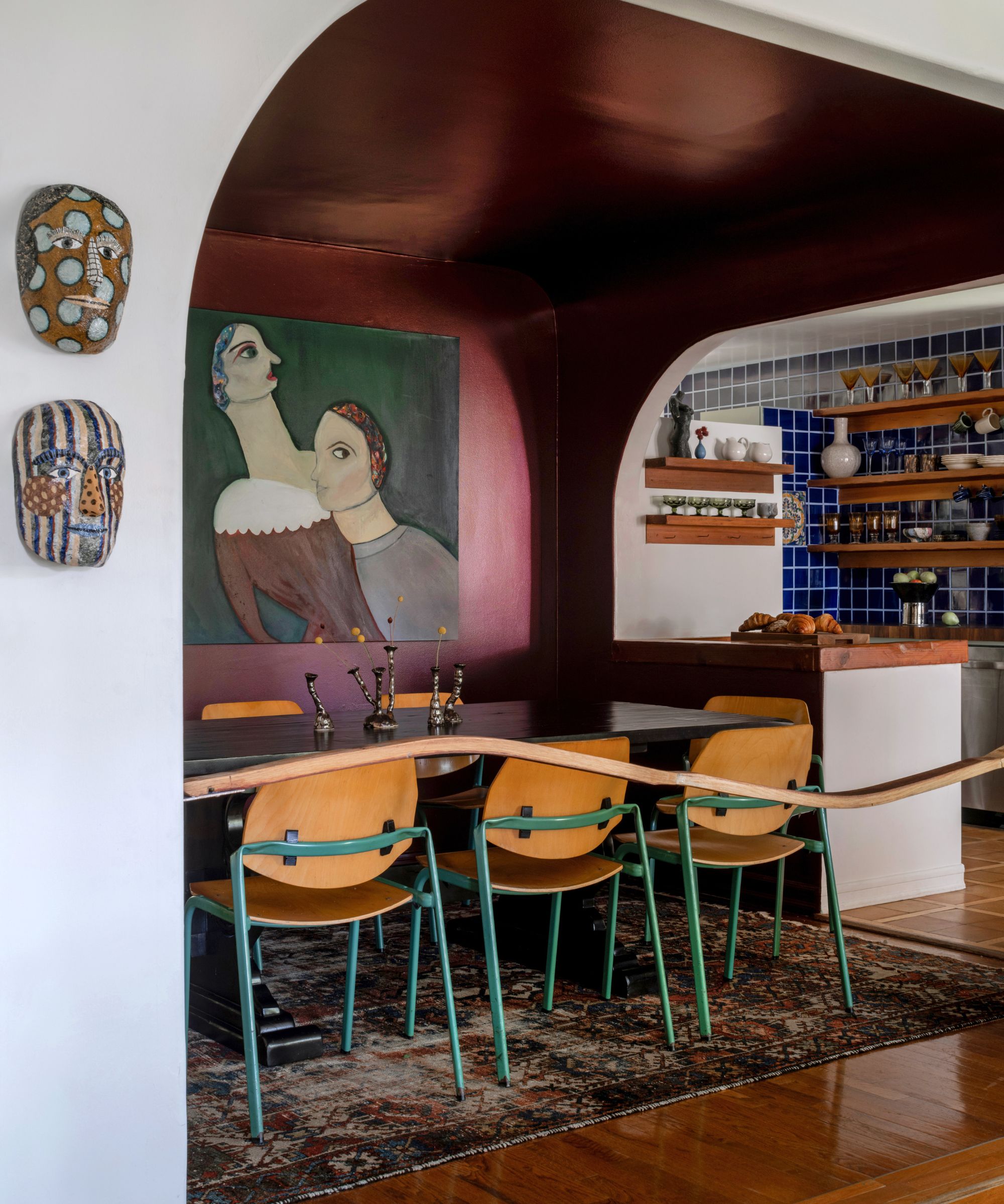 A dining room color-drenched with dark red, a large piece of artwork on the wall, a large black dining table with wooden chairs, and a patterned rug underneath.