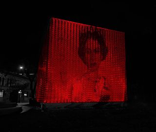 Image of Her Majesty The Queen projected on the side of a large metal cube in red light