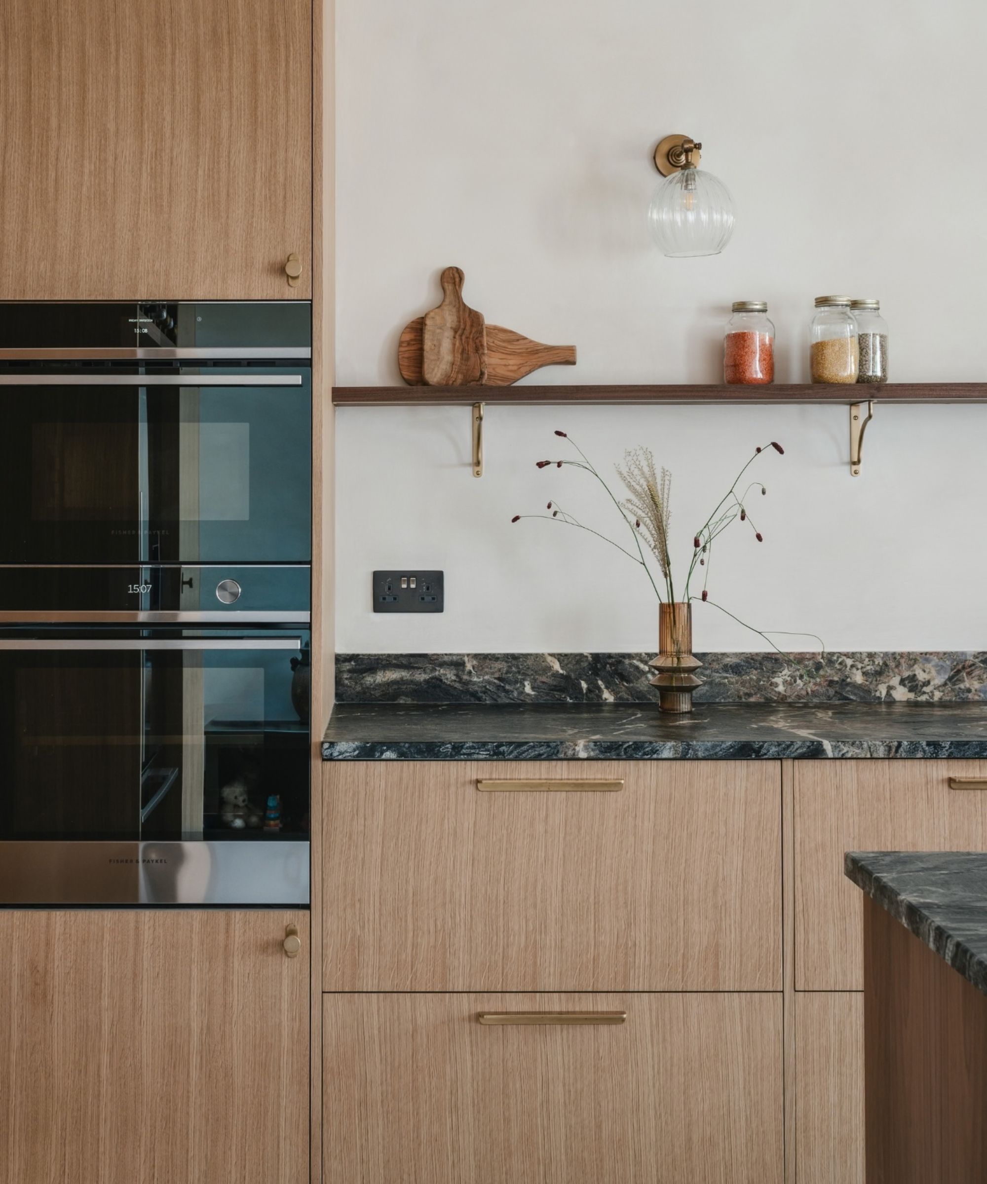 A modern kitchen featuring light oak wood cabinetry with brushed brass bar handles and round knob hardware. A stack of two built-in ovens is integrated into a tall cabinet on the left. The worktop is dark veined marble. Above the counter, a wooden shelf with brass bracket supports holds wooden chopping boards and three glass storage jars filled with colourful spices.