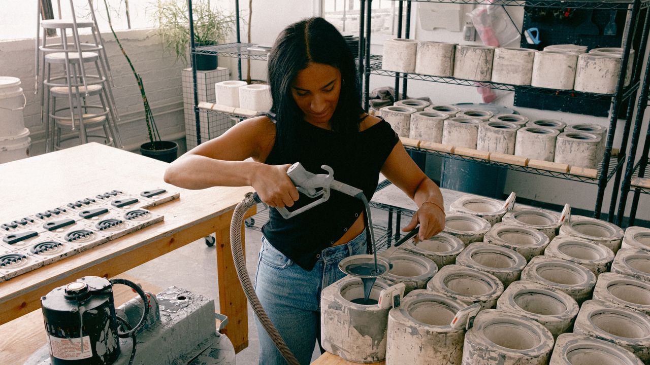 Image of a woman in a black shirt and jeans standing in a ceramics studio. She is filling glass-molds up with a dark gray liquid