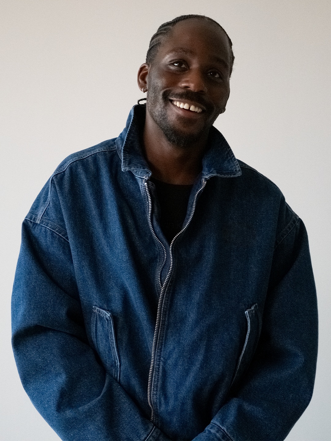 A young man poses in his industrial studio dressed in casual smartwear, sitting or standing in front of a gray wall with his sculptural creations.