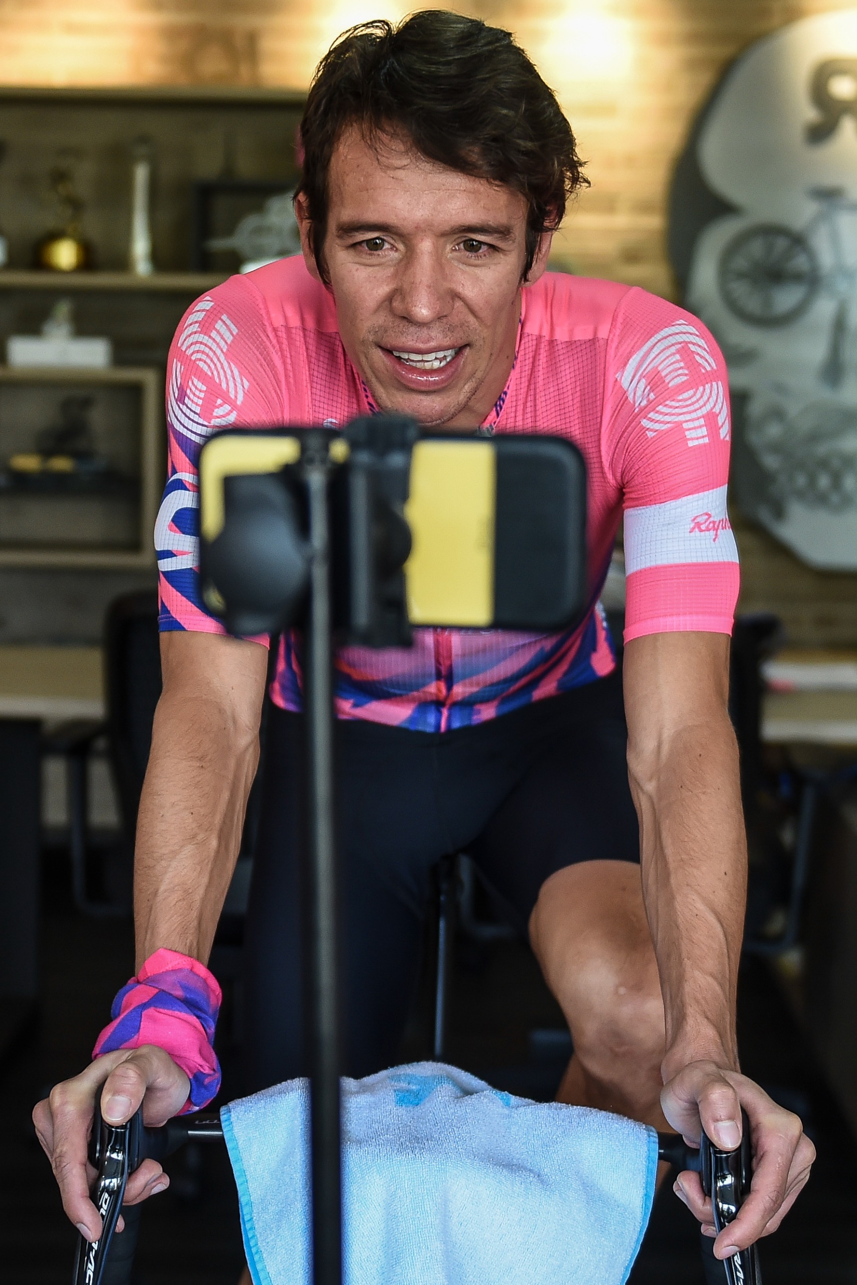 Rigoberto Uran of the Education First team records himself with a mobile phone as he rides a stationary bike while racing with fans through a virtual simulator
