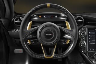 A close-up of the supercar's steering wheel in black with gold coloured shifters. The McLaren logo appears in the circular centre.