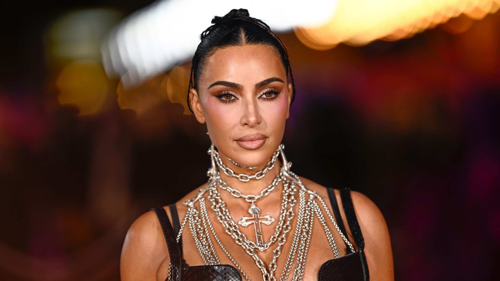 A woman with dark hair pulled back into a bun. She's wearing several silver chains around her neck and posing for a paparazzi photo on a red carpet.