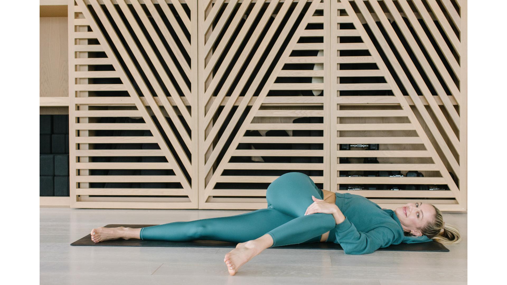woman dressed in jade green lying on her back performing a spinal twist with one bent knee crossing her body