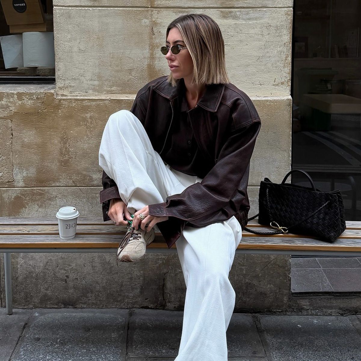 A woman wears a brown leather jacket, white sweatpants, and beige sneakers.