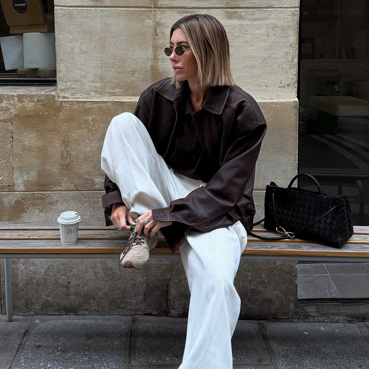 A woman wears a brown leather jacket, white sweatpants, and beige sneakers.