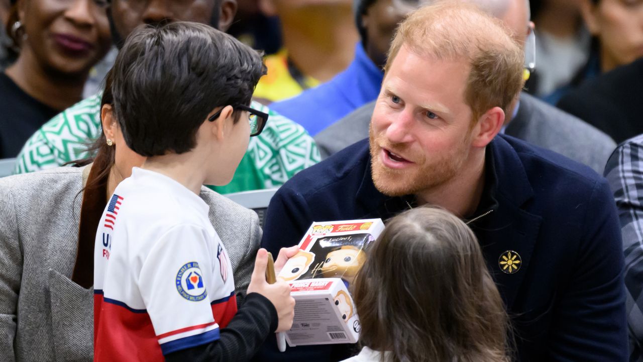 Prince Harry wearing a blue sweater talking ta. little boy holding a Prince Harry Funko Pop