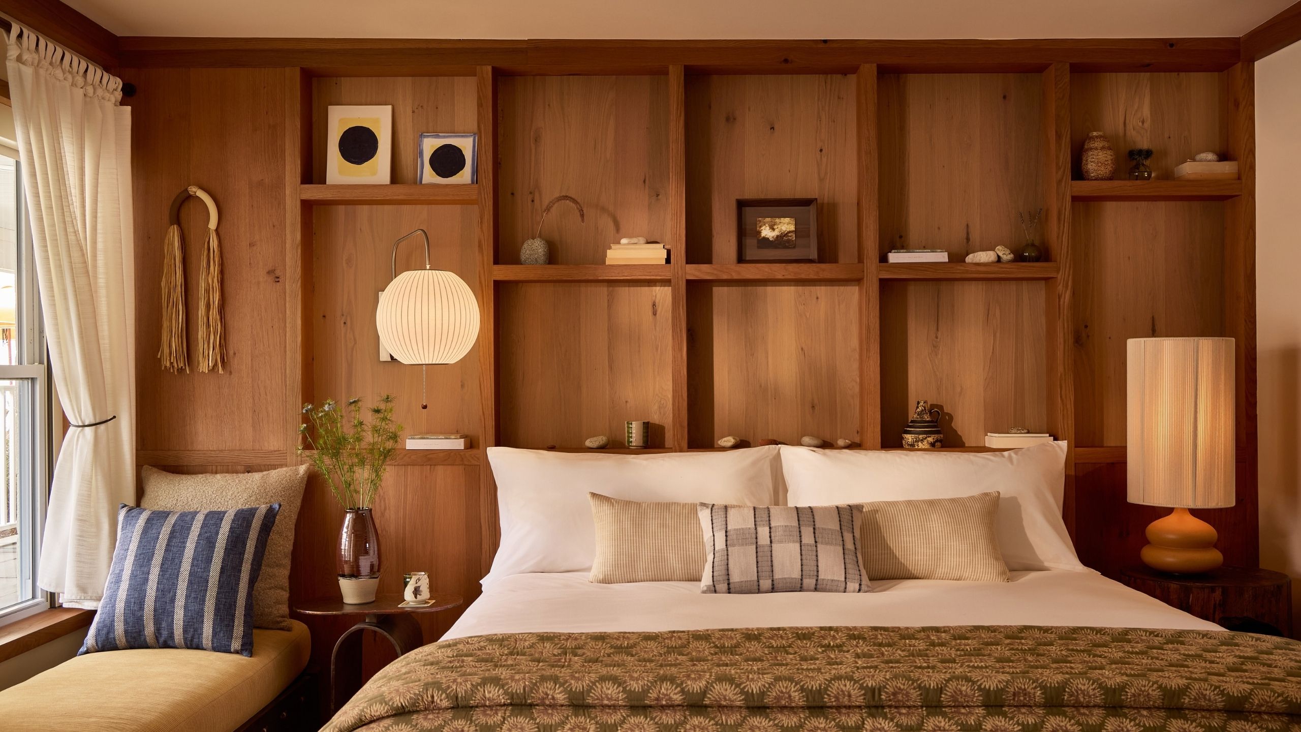 Interiors of a warmly furnished bedroom with ochre, brown, and terracotta tones, a king bed topped with an olive green runner, abstract artwork, warm lighting, and a window seat banquette.