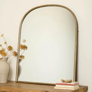 A brass arched mirror on a wooden table with a vase and books