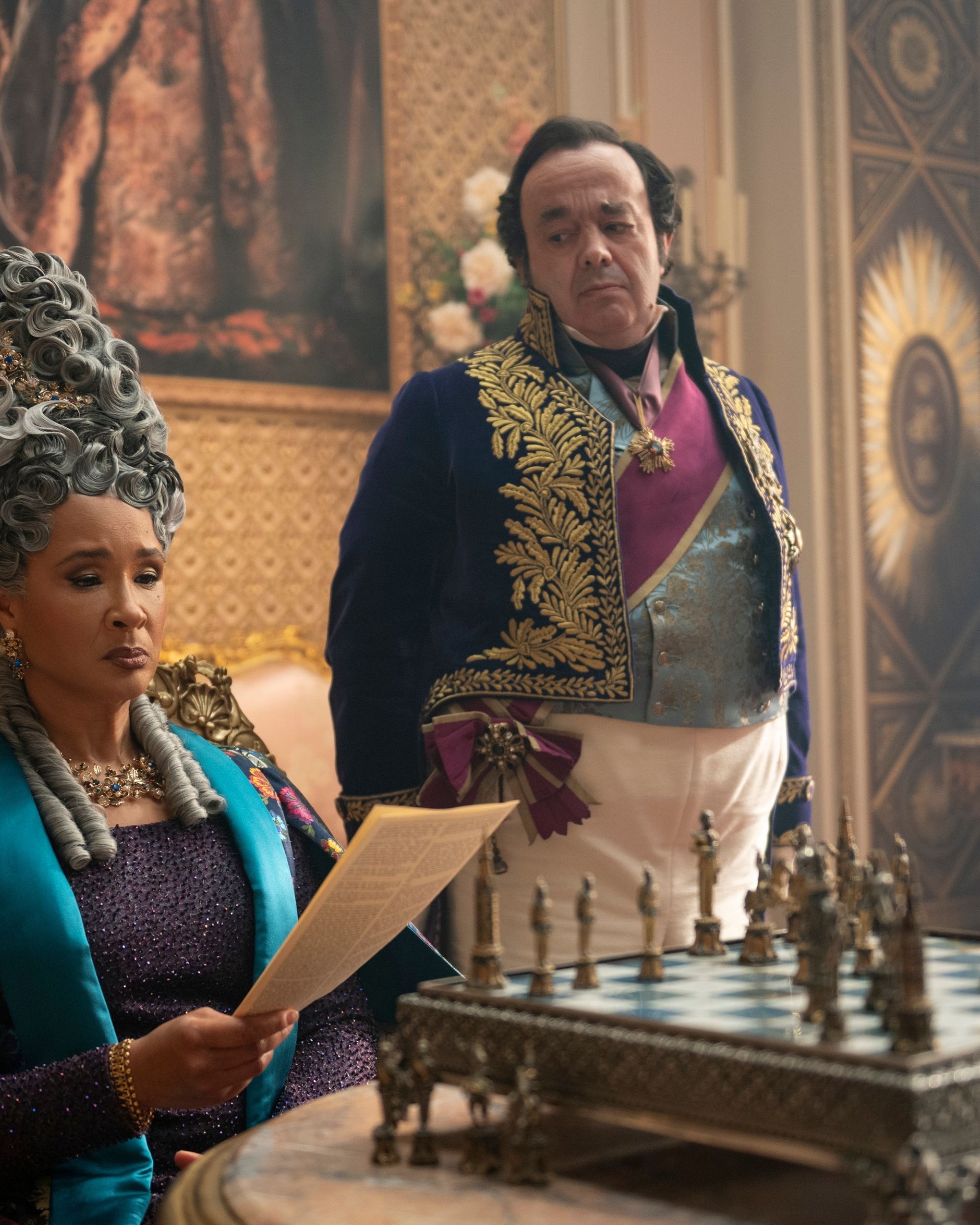 Production still from Netflix's Bridgerton series showing Queen Charlotte reading a paper in front of a lavish chessboard set.