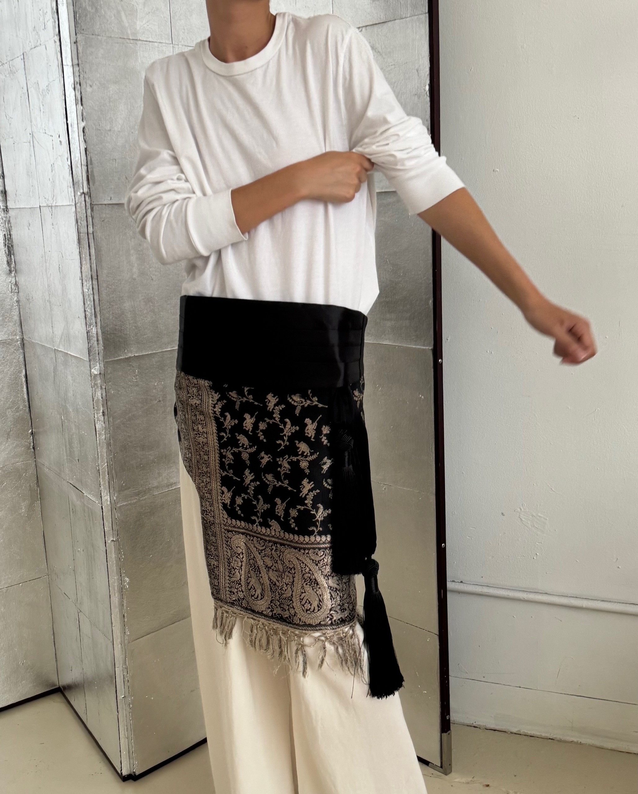 Andrea wears a white long-sleeve shirt, a black cummerbund, a paisley scarf, and beige trousers.