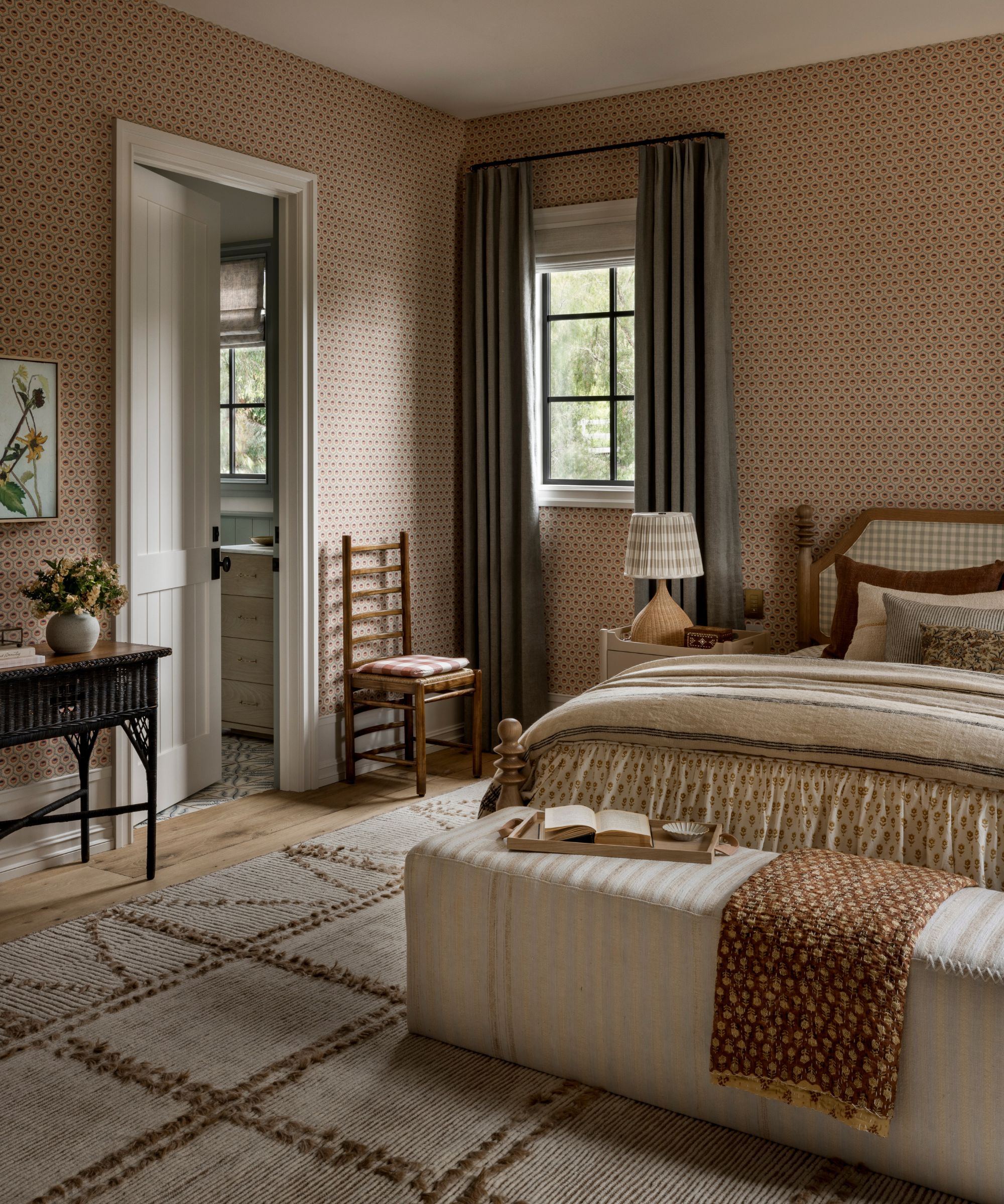 Country bedroom with ditsy wallpaper, gingham bed, beige rug and bedding, wooden vintage furniture, and window with long blue drapes.