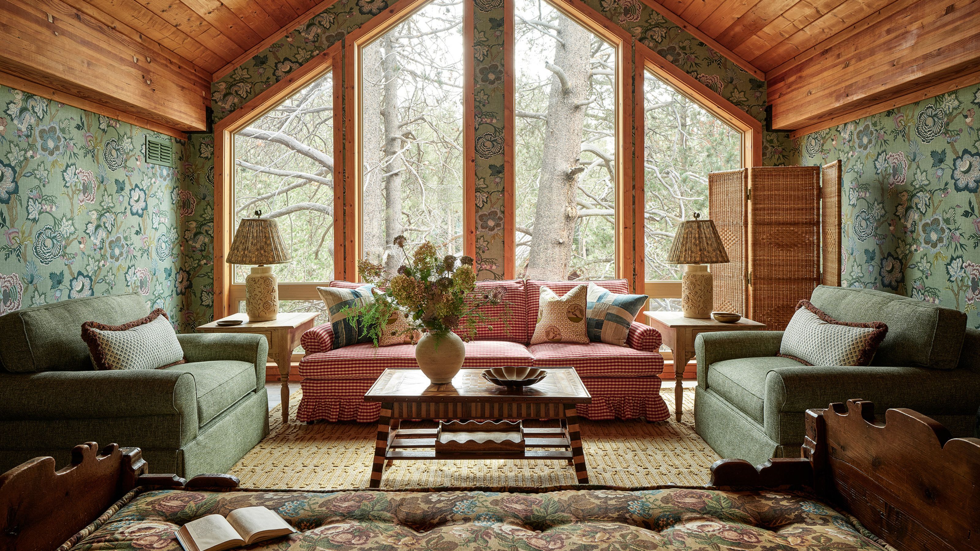 a living area with a vaulted wooden ceiling green floral wallpaper, large windows, gingham couch and two green armchaies with wicker and folk styling