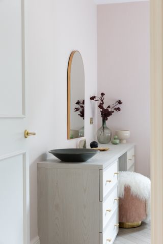 pale pink bedroom with pale grey vanity with drawers, sheepskin style stool, mirror hanging above, shallow bowl, vases and trinket holder on top