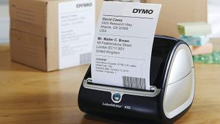 Address label being printed by DYMO LabelWriter 4XL thermal printer, on office desk in front of packages
