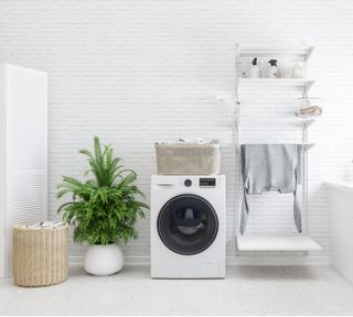 A white washing machine in a white tiled laundry room with woven laundry basket and indoor houseplant