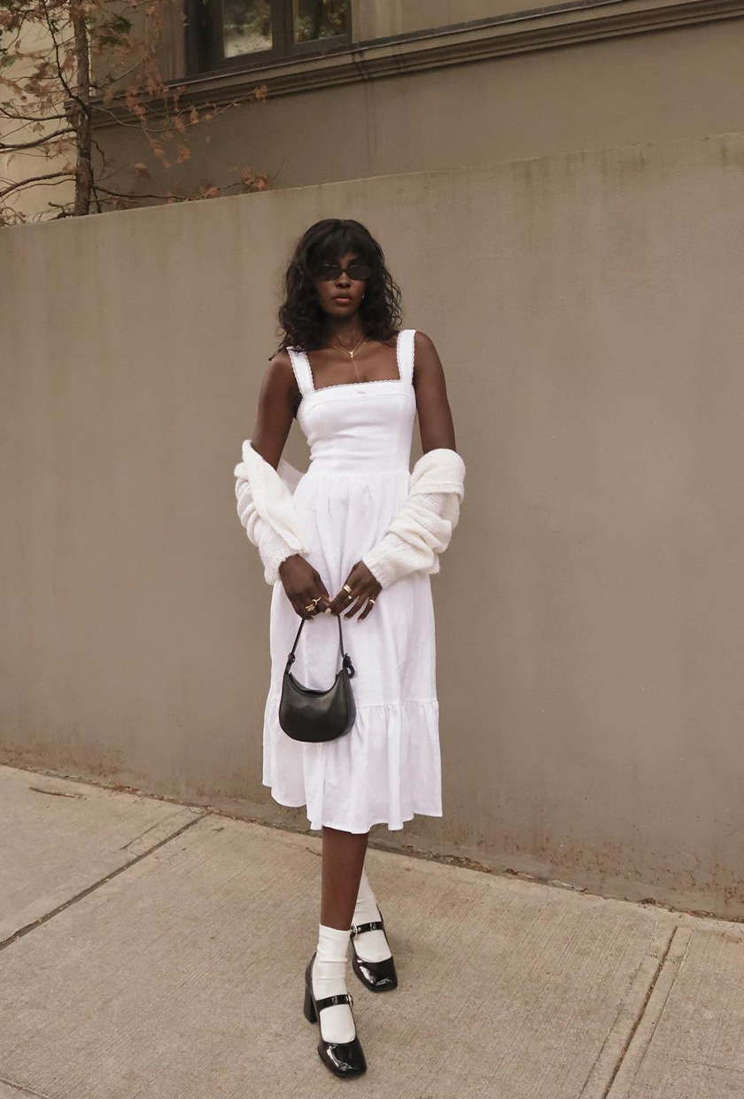 A woman's white dress outfit idea with a linen midi dress styled with a white cardigan, a black handbag, and black Mary Janes.