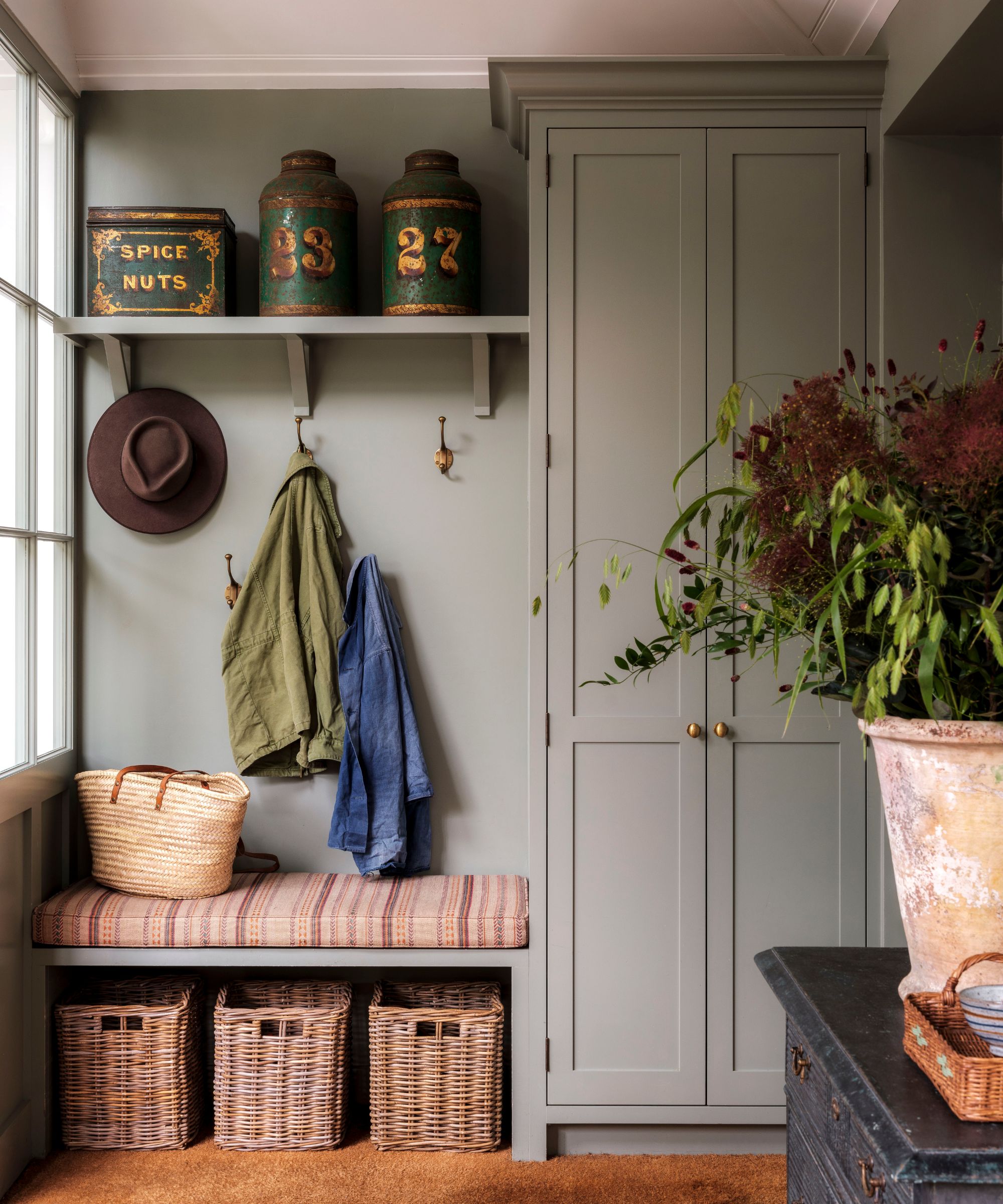 Entryway with grey walls and grey painted built-in cupboard, built-in bench with striped cushion, basket storage underneath and coats hanging on hooks, with vintage tin canisters on a shelf above