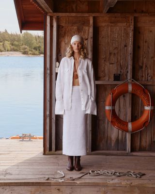 Elsa Hosk wearing a white headscarf, a white linen button down shirt, a white linen skirt, and brown boots.