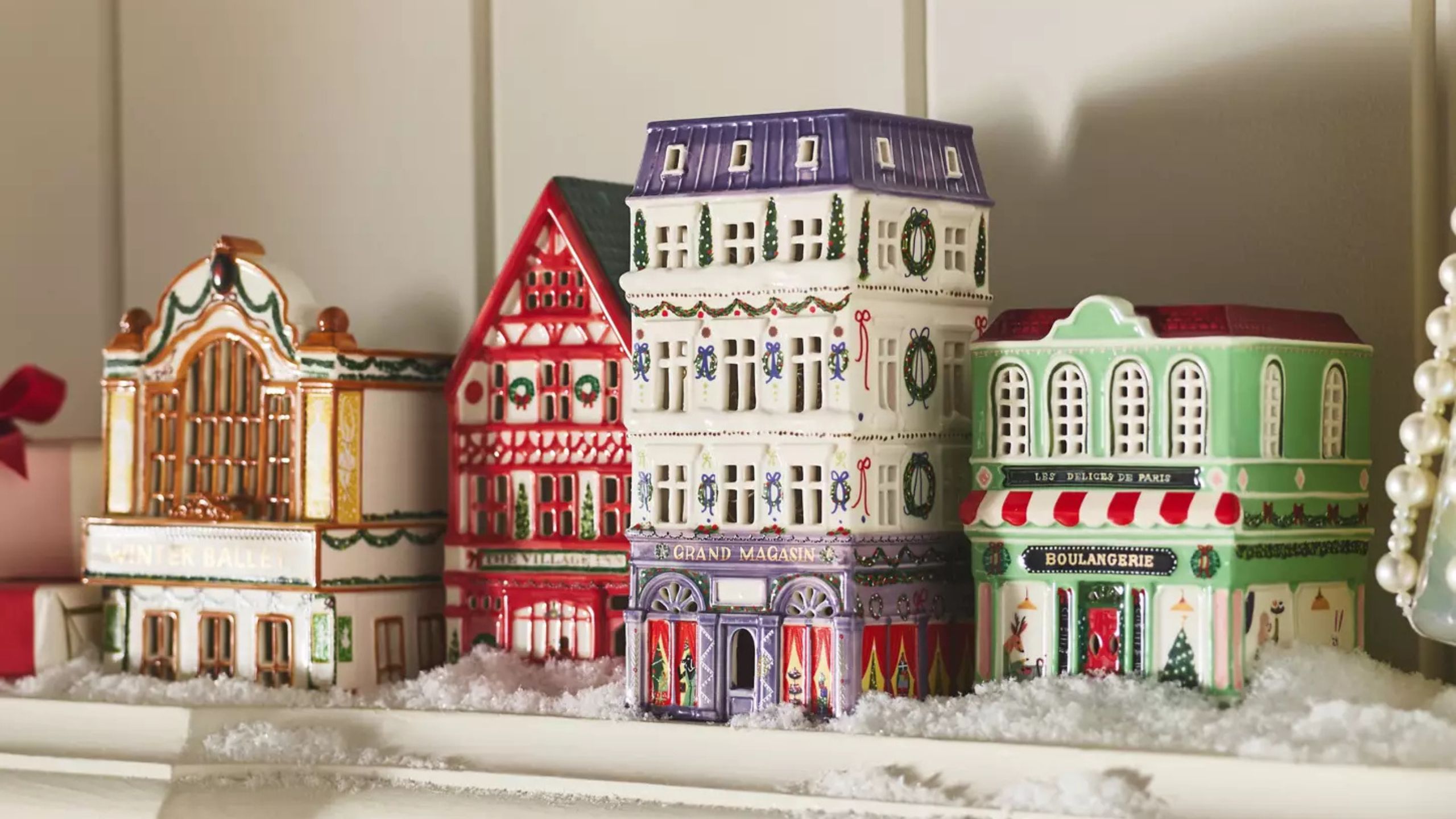 Four colorful ceramic Christmas houses sit on top of a mantelpiece in amongst fake snow and pearl decorations