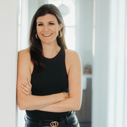 Image of a woman in a black top and pants leaning against a white wall.