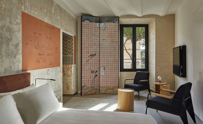 The Rooms of Rome bedroom with untreated concrete walls, bay window and trompe l’oeil panels that mimic a mirror