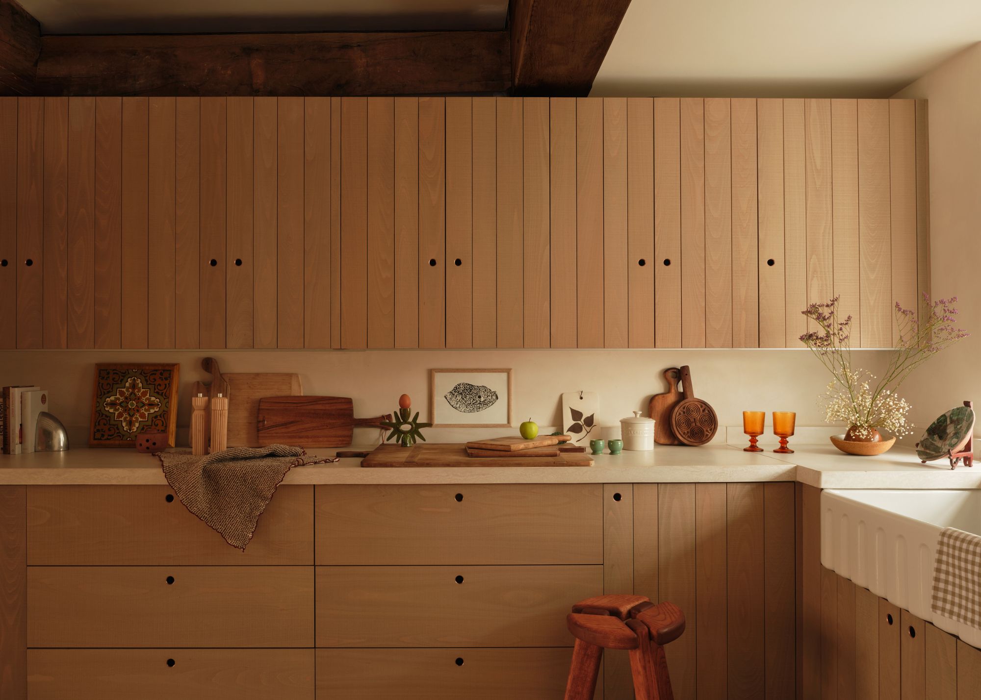 A wooden oak textured kitchen with upper and lower cabinets. On the kitchen countertops are wooden chopping boards, glassware plants and bowls. There is also other kitchen essentials scattered around. Beside the lower cabinet is a red small stool.