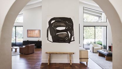 An archway-framed view of an open-plan living space with neutral walls and wooden floor. A black and white abstract artwork hangs on a wall in the centre