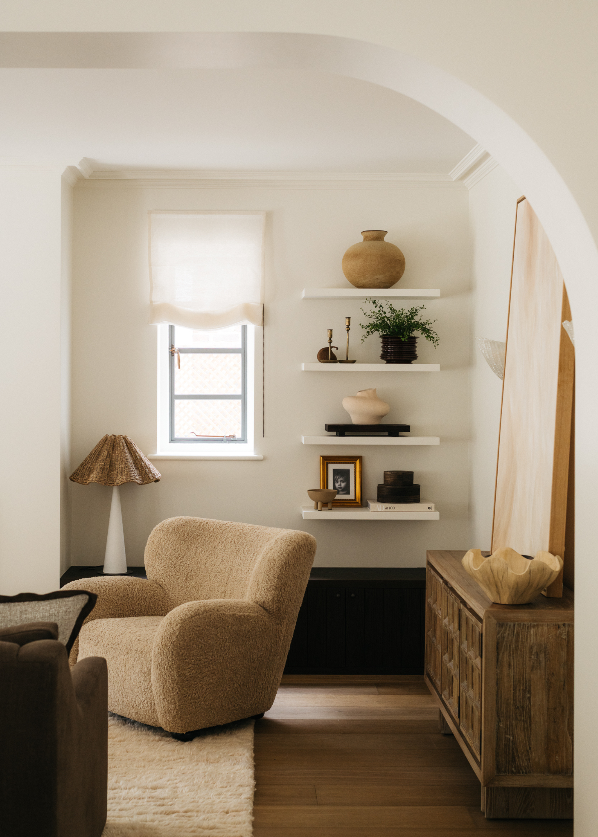 A neutral living room corner with an accent chair, a table lamp, open shelving with decorative objects and a console table with a piece of art resting on top