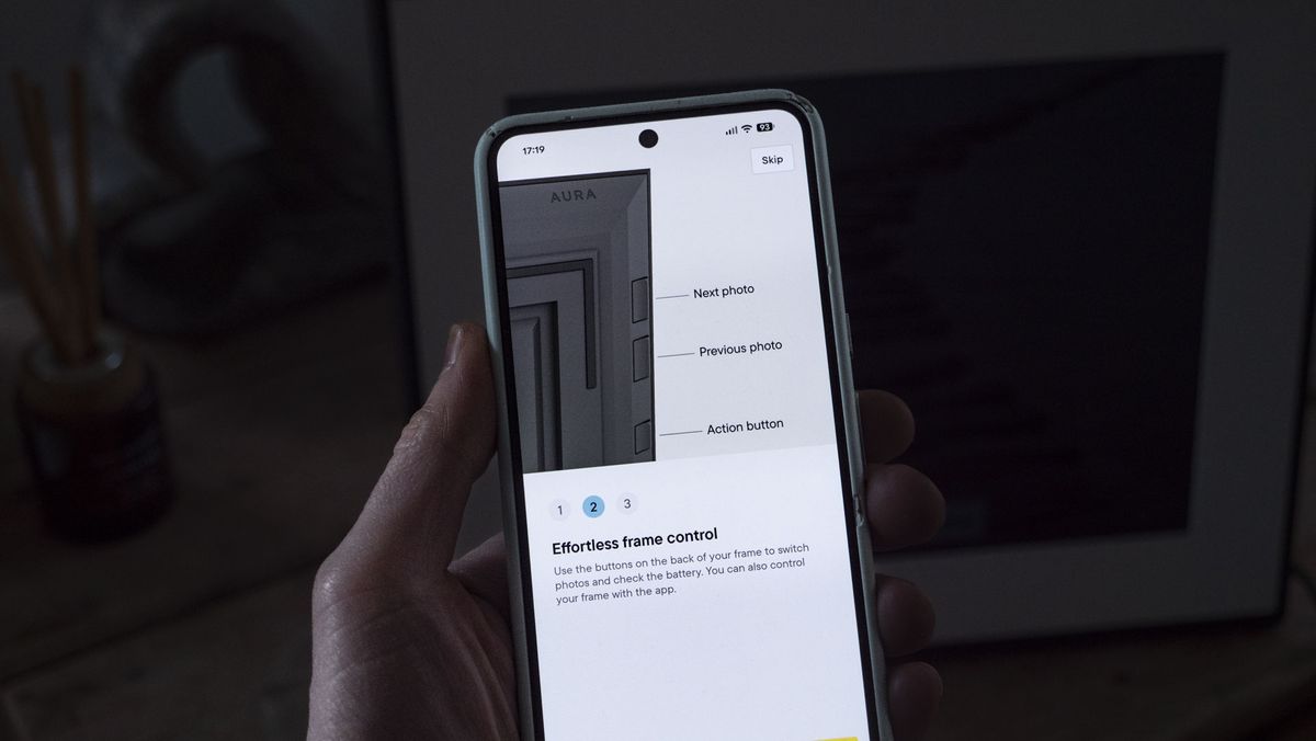 A phone being held, displaying the Aura companion app for digital photo frames, in front of the The Aura Ink 'E Ink' digital photo frame on a sideboard
