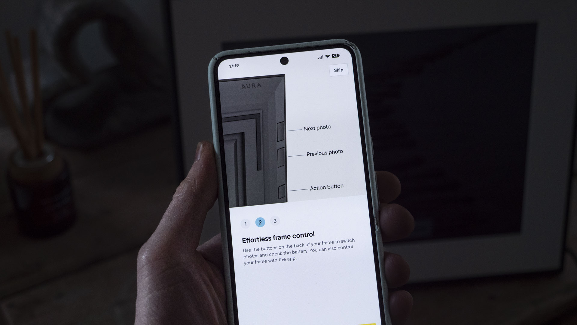 A phone being held, displaying the Aura companion app for digital photo frames, in front of the The Aura Ink 'E Ink' digital photo frame on a sideboard