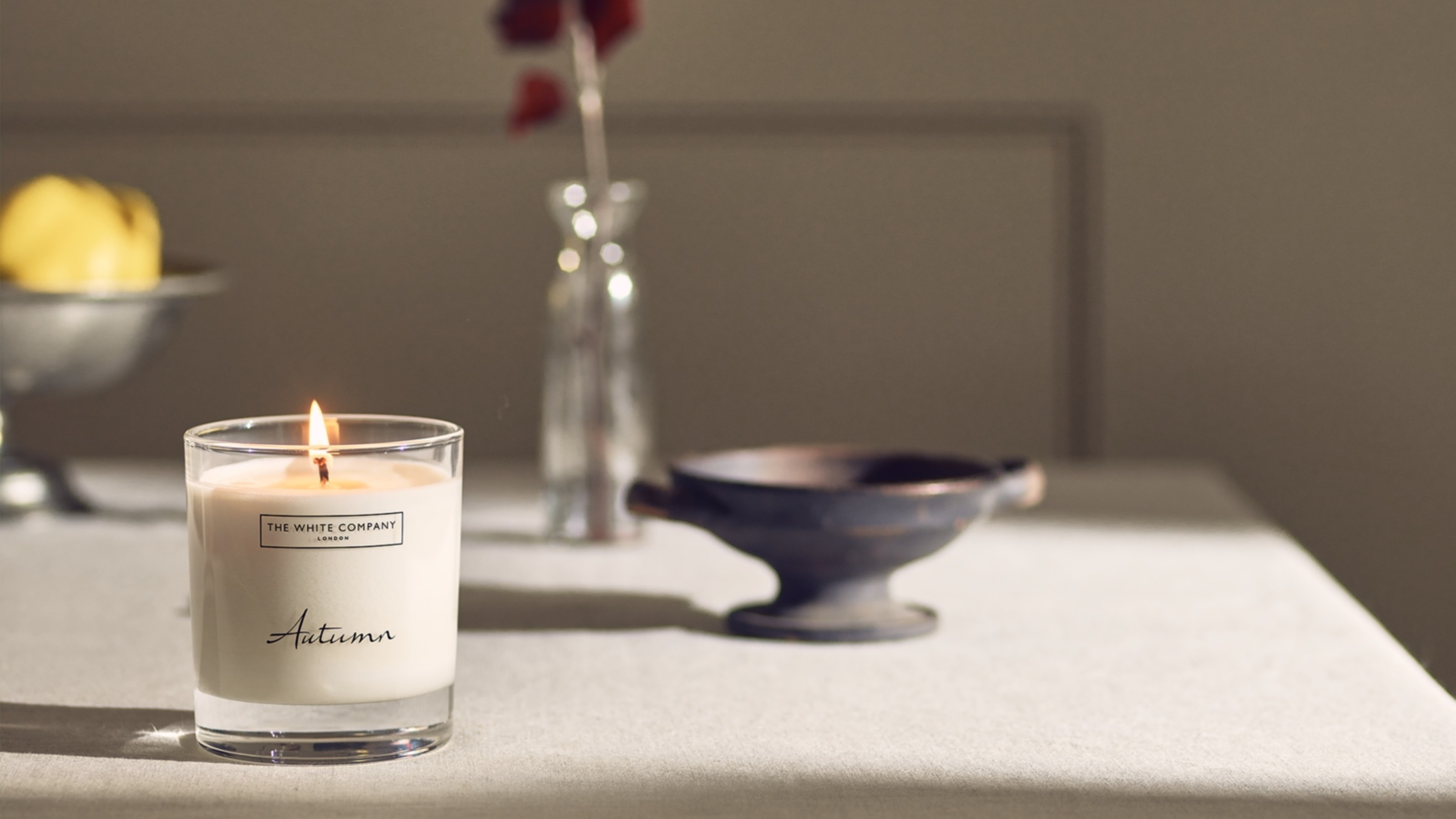 The White Company's lit Autumn signature candle on a dining table with a bowl of fruit and a vase with a single stem in the background