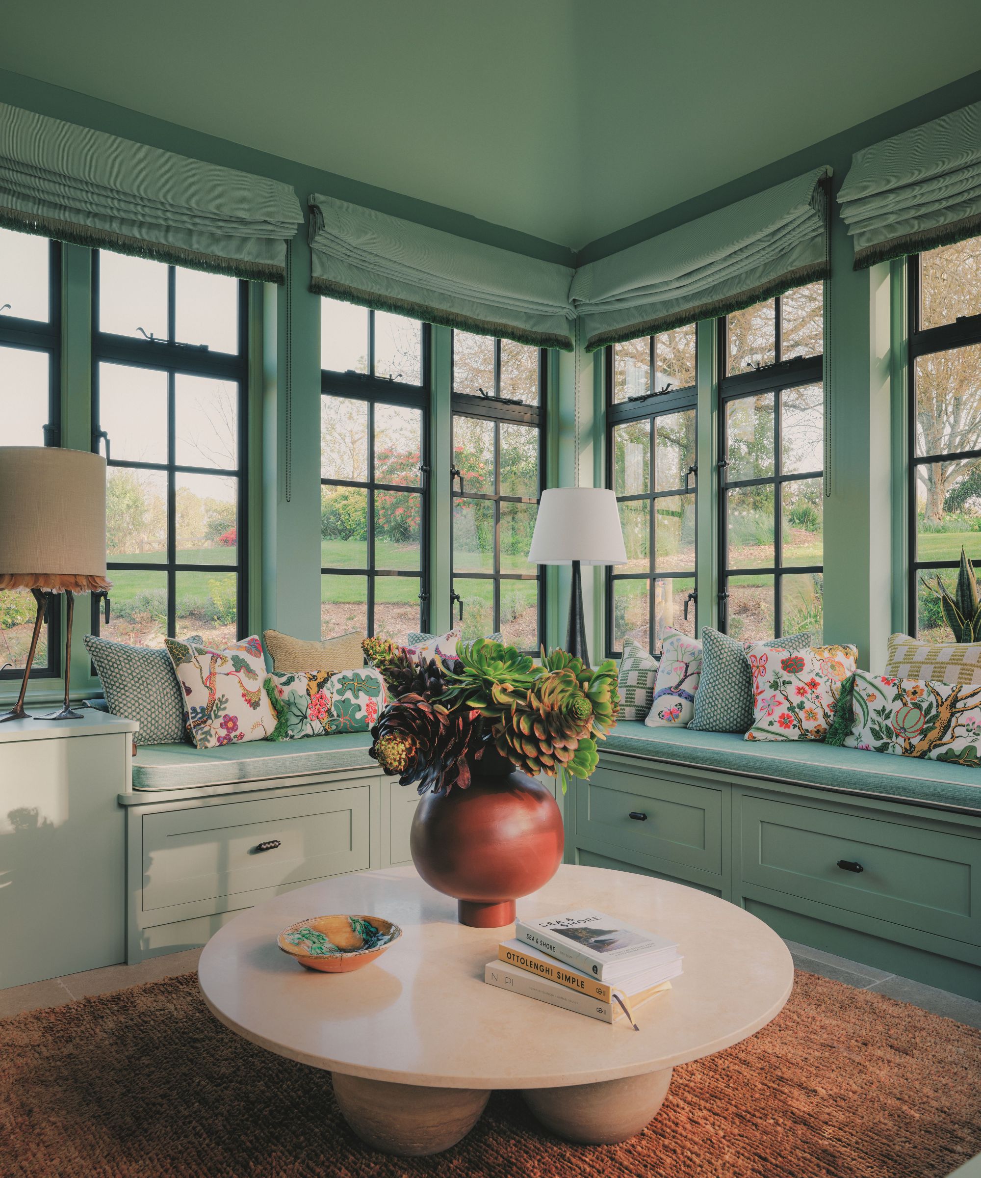Light-filled sunroom corner with green window frames, built-in bench seating with floral cushions, a beige round coffee table, and a terracotta succulent centerpiece.
