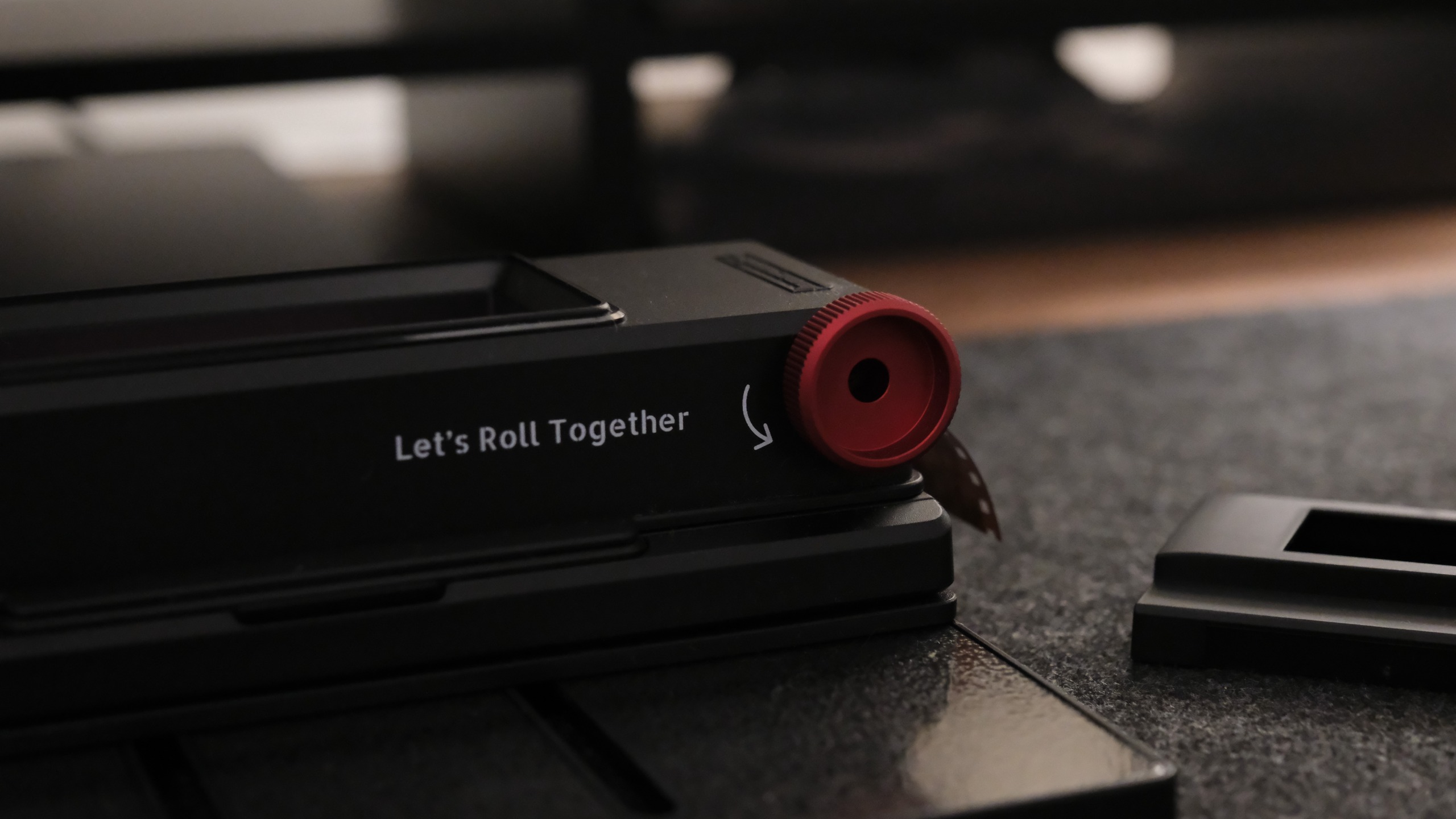 A macro shot focusing on the side of a black film scanning tool, highlighting a textured red dial next to white text that reads &amp;quot;Let's Roll Together&amp;quot; alongside a curved directional arrow.