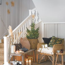 Hallway and staircase with a basket of wrapped presents, rattan rug, chairs and small decorated Christmas Tree with small table with lit candles.