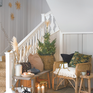 Hallway and staircase with a basket of wrapped presents, rattan rug, chairs and small decorated Christmas Tree with small table with lit candles.