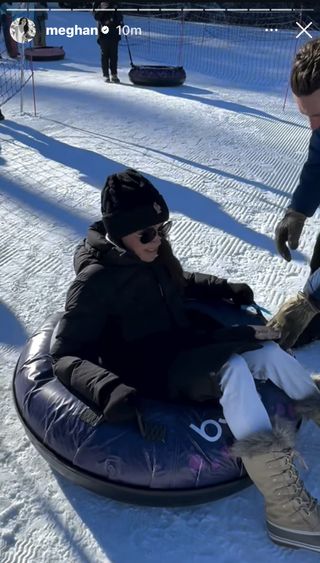 Meghan Markle wearing a black coat and white pants with fur boots sitting in a snow tube