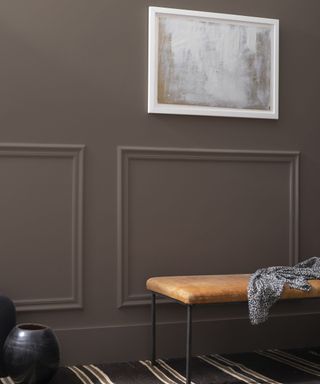 dark brown panelled wall with white artwork in white frame, tan bench with item of clothing draped