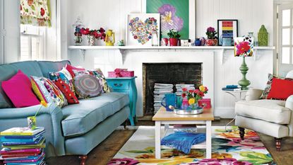 White living room, blue sofa with bright multicoloured cushions, bold floral rug, wooden coffee table, white sofa, stacked books used as table, stacked magazines in fireplace, wooden floorboards, 3D heart art on shelf, potted house plants.