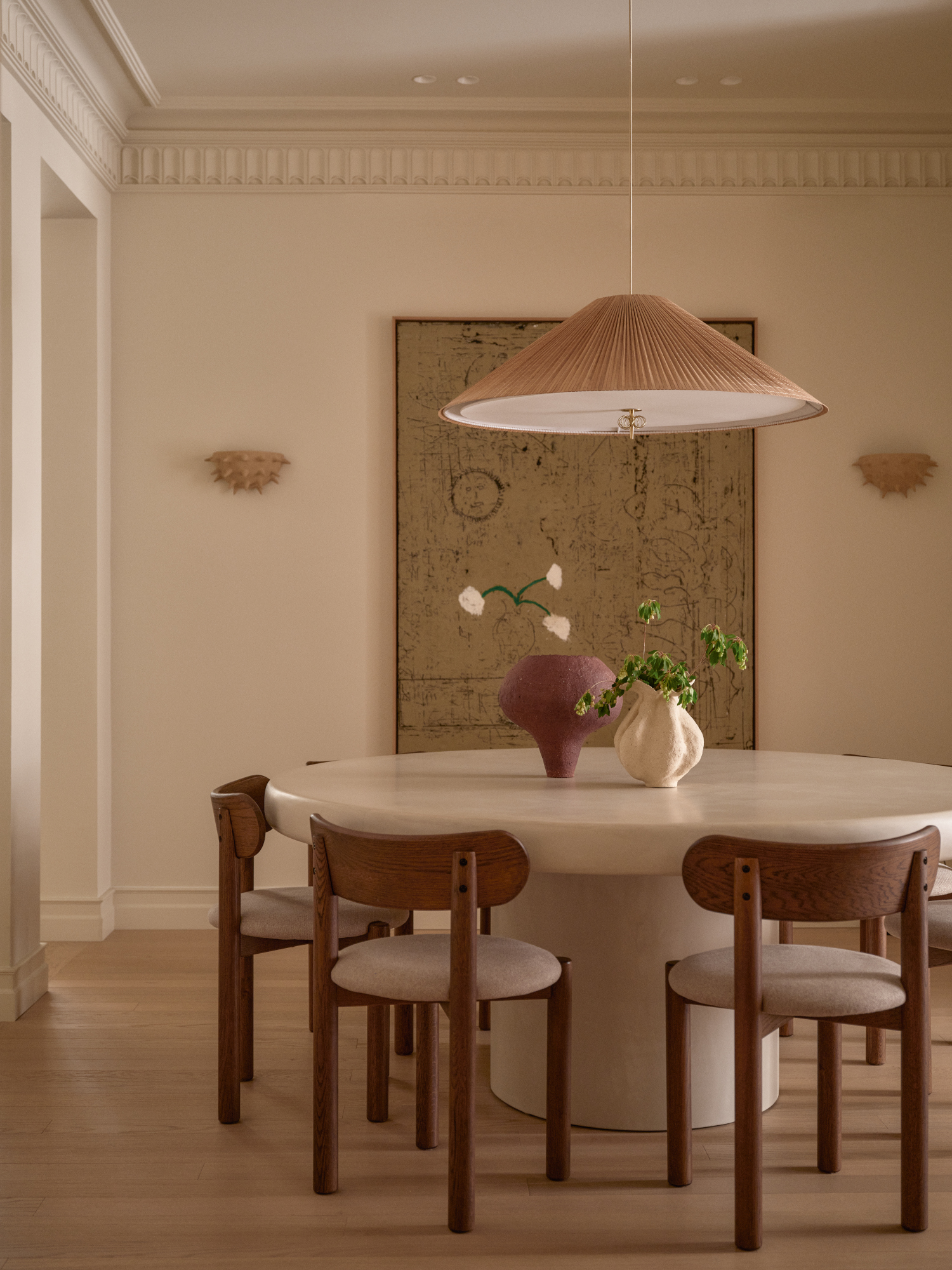 Warm white dining room with round microcement table, wood and cream chairs, pleated pendant and burgundy and cream vases