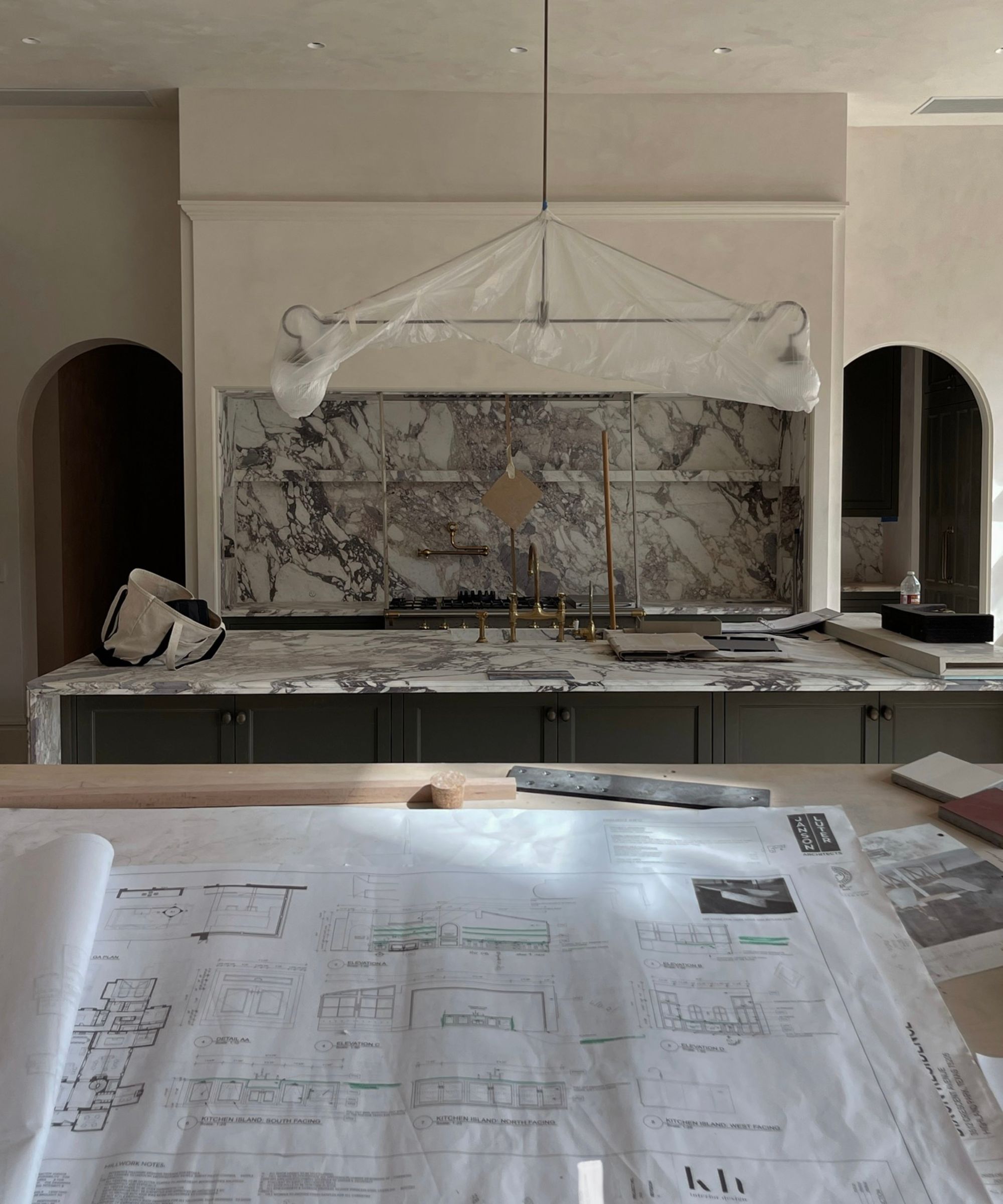 Image of modern kitchen under construction, plans of design in foreground, with large island, marble backsplash and lighting in background