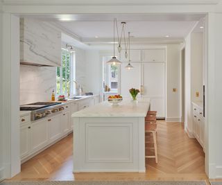 A white kitchen with marble countertops, backsplash, and range hood. A kitchen island sits in the middle with a fruit bowl and a glass vase of flowers to decorate. Natural wood counter stools with woven seats add bar seating and ties n with the wood herringbone floor.