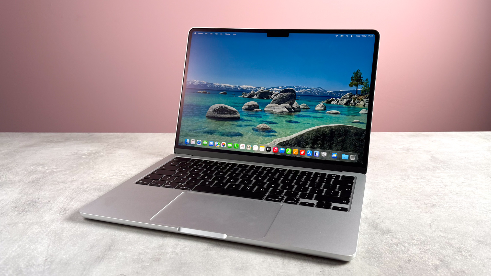 The MacBook Air M5 open and powered on on a stone effect surface in front of a pink background.