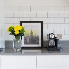 A white kitchen with a vase of flowers, a framed art print and a pod coffee machine