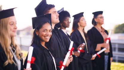 A cheerful group of graduates wearing caps and gowns stand in a line, each holding a diploma tied with a red ribbon
